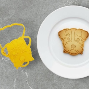Puede incluir: Un cortador de galletas amarillo con forma de perro junto a una galleta con forma de perro en un plato blanco. La galleta es de color marrón claro con detalles faciales. El cortador es de plástico flexible.