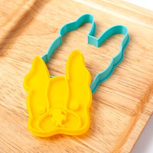 May include: A yellow plastic cookie cutter in the shape of a French Bulldog's face, with a blue outline. The cutter is on a wooden surface.
