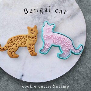 Può includere: Un biscotto a forma di gatto del Bengala e un tagliabiscotti rosa con contorno turchese, entrambi a forma di gatto del Bengala. Il biscotto è marrone chiaro con macchie marroni scure. Il testo "Bengal cat" e "cookie cutter&stamp" è nell'immagine.