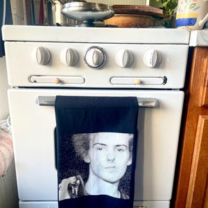 May include: A black and white kitchen towel with a portrait of a person hangs on a white oven. The oven has five white knobs and a silver handle. A stainless steel pan sits on top of the stove.