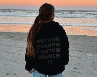 May include: A person wearing a black hoodie with the text "NOTHING CHANGES IF NOTHING CHANGES!!!" on the back, standing on a sandy beach with the ocean in the background.