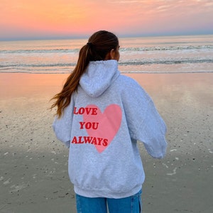 May include: A person wearing a gray hoodie with a pink heart and the words "LOVE YOU ALWAYS" on the back, standing on a beach with a sunset in the background.