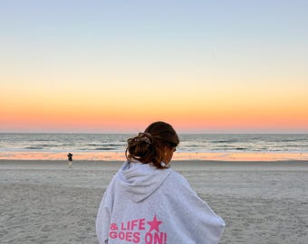 May include: A person wearing a gray hoodie with the text "& LIFE GOES ON!" in pink lettering and a pink star on a beach at sunset.