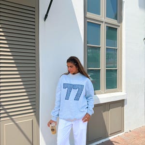 May include: A light gray sweatshirt with the number "77" in navy blue stripes. The sweatshirt is paired with white pants. The person is holding a clear plastic cup with a brown beverage.
