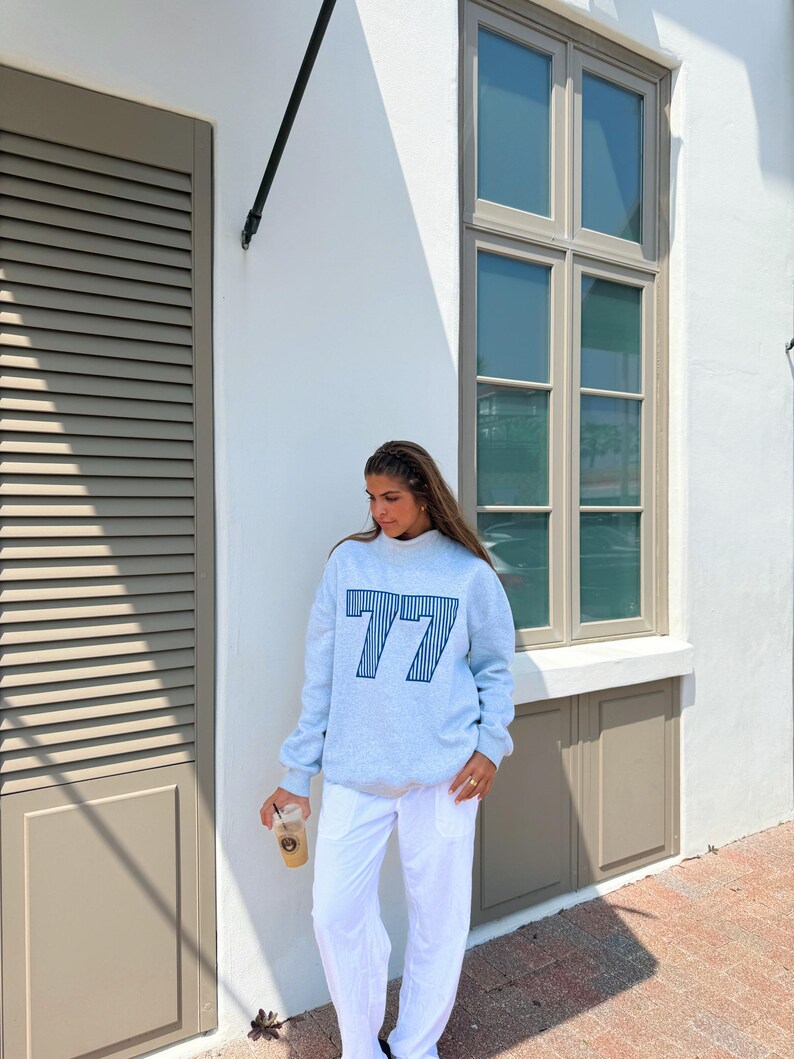 May include: A light gray sweatshirt with the number "77" in navy blue stripes. The sweatshirt is paired with white pants. The person is holding a clear plastic cup with a brown beverage.