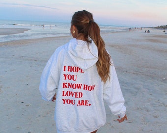 May include: A person wearing a gray hoodie with the text "I hope you know how loved you are." in red lettering. The person is walking on a sandy beach.