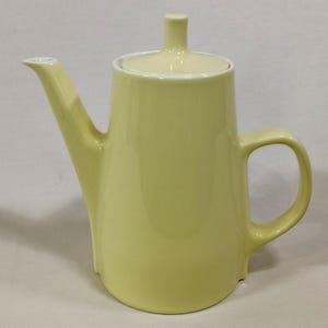 Puede incluir: Una tetera de cerámica amarillo pálido con tapa y asa a juego. La tetera tiene un pico largo y un cuerpo redondeado. La tapa tiene un pequeño pomo cilíndrico. La tetera probablemente se usa para preparar té.