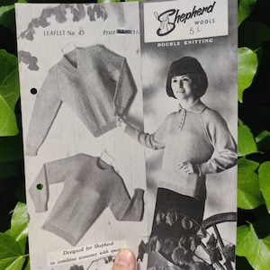Puede incluir: Folleto de patrones de tejido vintage con una mujer modelando un suéter con cuello y ilustraciones de otros diseños. El folleto "Shepherd Wools" incluye el texto "Double Knitting". El folleto se sostiene contra un fondo de hojas verdes.