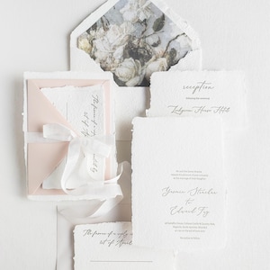 Könnte beinhalten: Ein Hochzeitseinladungs-Set aus handgeschöpftem Papier mit Büttenrand, einem hellrosa Umschlagfutter und einem floral gemusterten Umschlaginneren. Das Set enthält eine Einladung, eine Empfangskarte und eine Antwortkarte, zusammengebunden mit einem weißen Band.