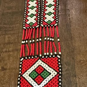 May include: A long beaded necklace featuring a rectangular pendant and two smaller rectangular sections connected by multiple red beaded strands. The design incorporates a geometric pattern in red, white, green, and black beads with a fringe of green beads.
