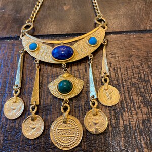 May include: A gold necklace with a crescent moon pendant adorned with blue and green stones. The pendant has five dangling coins with intricate designs.