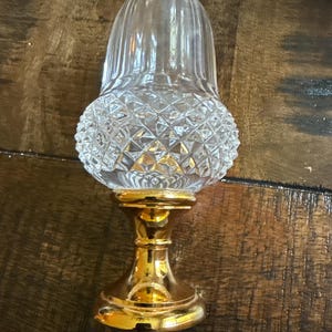 Puede incluir: Una pantalla de lámpara de vidrio transparente con un patrón de diamantes en la mitad inferior y líneas verticales en la parte superior. La pantalla está montada sobre una base de metal dorado. La pantalla de la lámpara está diseñada para colocarse sobre una mesa.