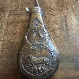 Op de afbeelding: Een vintage koperen kruitfles met reliëfversieringen. Het bovenste gedeelte toont twee vogels, terwijl het onderste gedeelte een hond in een veld weergeeft. De fles heeft een taps toelopende vorm met een metalen tuit en een donkere patina.