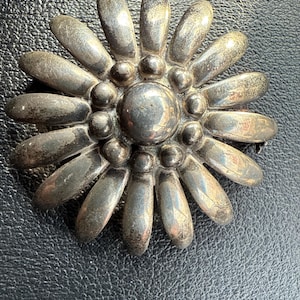 Może przedstawiać: Srebrna metalowa broszka w kształcie kwiatu. Centralny okrąg otoczony jest płatkami. Broszka ma wygląd vintage, z lekko przyciemnionym wykończeniem. Płatki i centralny wzór są szczegółowe z małymi, wypukłymi okrągłymi akcentami.