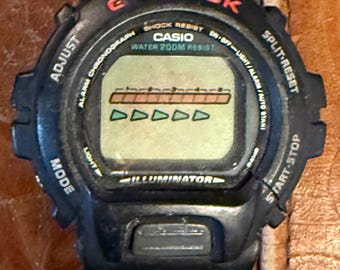 Puede incluir: Reloj digital G-Shock negro con texto rojo y blanco. La esfera muestra líneas horizontales y triángulos. Botones para ajustar, modo, luz y inicio/parada. Resistente al agua.