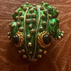 Puede incluir: Broche verde con forma de rana con detalles dorados y ojos de piedra negra. El broche tiene una superficie texturizada con protuberancias doradas y líneas doradas. Los ojos de la rana están engastados en marcos dorados.
