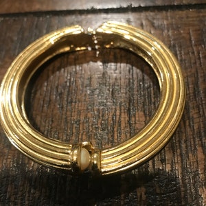 Puede incluir: Un brazalete dorado con un acento ovalado blanco. El brazalete tiene un diseño acanalado.