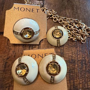 May include: A set of Monet clip-on earrings and a necklace. The earrings are white with a gold-colored center stone and a gold-colored border. The necklace has a gold-colored chain. The earrings are on a brown card with the word "MONET" printed on it.