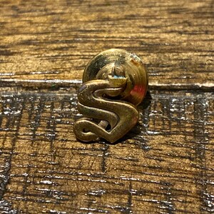 Op de afbeelding: Een gouden metalen pin met een gestileerd letter 'S'-ontwerp. De pin heeft een ronde achterkant met een puntige punt.