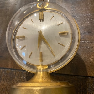 May include: A gold-toned table clock with a clear glass dome and a white face. The clock face has black Roman numeral hour markers and black hands. The clock face also has the word "GUBELIN" printed on it.