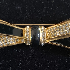 Könnte beinhalten: Eine goldfarbene Schleifenbrosche mit schwarzem Emaille und klaren Strasssteinen.