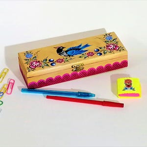 May include: A wooden box with a bird and floral design, a gold clasp, and a pink scalloped edge. The box is surrounded by pens, paperclips, and a yellow eraser with a floral design.