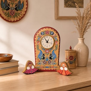 May include: A wooden clock with a floral and rooster design, featuring a round clock face. Two wooden figurines of women in traditional dress are in front of the clock. A wooden plate and a small jar are also visible.