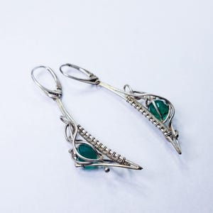 Può includere: Un paio di orecchini in argento con chiusura a leva. Ogni orecchino presenta una pietra preziosa verde smeraldo a forma di goccia, incastonata in un design avvolto in filo metallico. Gli orecchini hanno un'estetica moderna e geometrica.