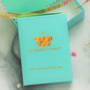 Puede incluir: Una caja de regalo turquesa con un logotipo dorado y el texto "CHARMED COAST" y "www.charmedcoast.com". La caja est&aacute; abierta, revelando un vistazo del interior. Cinta dorada y acentos florales visibles.