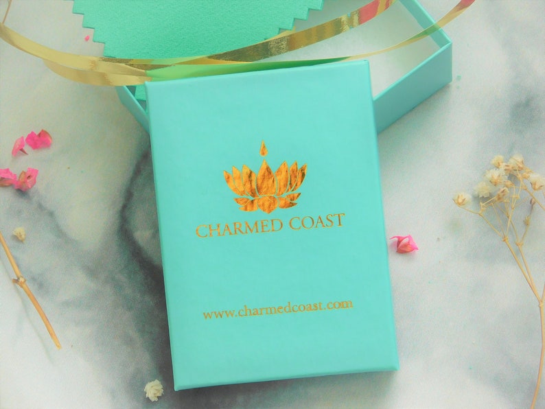 Puede incluir: Una caja de regalo turquesa con un logotipo dorado y el texto "CHARMED COAST" y "www.charmedcoast.com". La caja est&aacute; abierta, revelando un vistazo del interior. Cinta dorada y acentos florales visibles.
