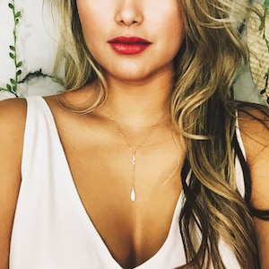 May include: A gold chain necklace with a teardrop-shaped pendant. The pendant is white and has a subtle sparkle. The necklace is worn by a person with long blonde hair.