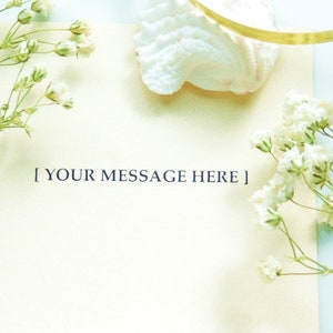 K&ouml;nnte beinhalten: Eine leere elfenbeinfarbene Karte mit dem Text "[ YOUR MESSAGE HERE ]" in dunkelblau gedruckt. Die Karte ist von wei&szlig;en Blumen und einer wei&szlig;en Muschel umgeben.