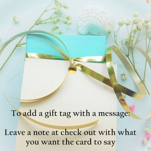 Puede incluir: Una caja de regalo turquesa con una tarjeta blanca, adornada con una cinta dorada. La tarjeta dice: "Para agregar una etiqueta de regalo con un mensaje: Deja una nota al finalizar la compra con lo que quieres decir en la tarjeta."
