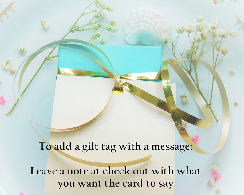 Puede incluir: Una caja de regalo turquesa con una tarjeta blanca, adornada con una cinta dorada. La tarjeta dice: "Para agregar una etiqueta de regalo con un mensaje: Deja una nota al finalizar la compra con lo que quieres decir en la tarjeta."