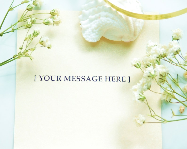 Puede incluir: Una tarjeta color crema con el texto "[ YOUR MESSAGE HERE ]" est&aacute; rodeada de flores blancas y una concha sobre un fondo azul claro. Una cinta dorada es visible cerca de la concha. La imagen tiene una est&eacute;tica suave y aireada.