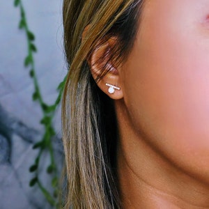 Puede incluir: Primer plano de una oreja adornada con dos pendientes. Uno es una peque&ntilde;a gema blanca y redonda. El otro es una barra horizontal con peque&ntilde;as gemas transparentes. El fondo es una suave mezcla borrosa de colores.