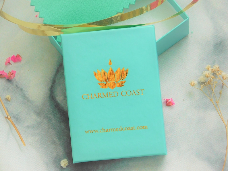 K&ouml;nnte beinhalten: Eine t&uuml;rkisfarbene Geschenkbox mit einem goldenen Logo, auf dem "CHARMED COAST" und die Website-Adresse "www.charmedcoast.com" steht.