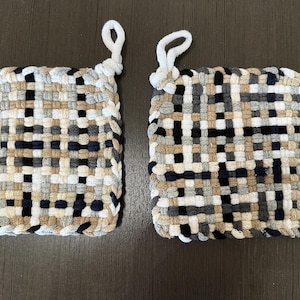 May include: Two square woven potholders with a loop for hanging. The potholders are made of woven fabric in a pattern of black, white, gray, and tan. The potholders are on a dark brown surface.