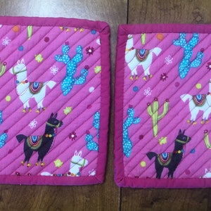 May include: Two pink quilted pot holders with a llama and cactus pattern. The pot holders have a pink border.