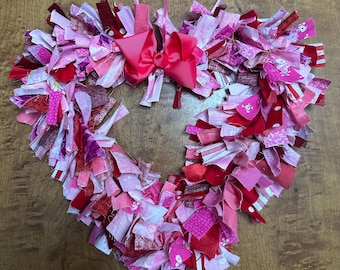May include: A heart-shaped wreath made of pink, red, and white fabric scraps. The wreath is decorated with a large pink bow.
