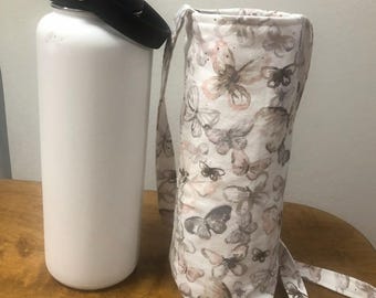 May include: A white insulated water bottle with a black lid and handle sits next to a fabric water bottle carrier. The carrier is white with a pattern of butterflies in shades of brown and pink. The carrier has a matching strap.