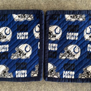 May include: Two quilted rectangular placemats. The placemats are navy blue with a repeating pattern of white American football helmets and the word "COLTS" in white. The edges are trimmed with a navy blue binding.