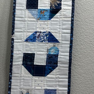 May include: A white fabric wall hanging with a blue and white patchwork design spelling out the word "JOY". The fabric is quilted and has a blue and white star border.