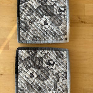 May include: Two square quilted pot holders. The fabric features a black and white farm-themed print with text and illustrations of produce, including corn, tomatoes, and peppers. The edges are trimmed with grey fabric.