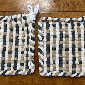 May include: Two square potholders featuring a woven grid pattern in shades of white, charcoal grey, and tan. Each potholder has a thick, braided border and a small loop at one corner for hanging, constructed from soft, textured fabric strips.
