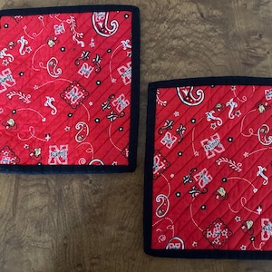 May include: Two square red quilted pot holders with a black border. The fabric features a paisley and floral pattern with the letter "N" in a square design. The pot holders are on a wooden surface.