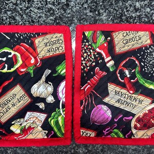 May include: Two red and black quilted pot holders with a Cajun Creole theme. The pot holders feature images of crawfish, peppers, garlic, and other spices. The text on the pot holders reads "Louisiana Crawfish", "Hot N Spicy", "Cajun Creole", and "Jambalaya Gumbo".