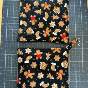 May include: Two square fabric pot holders featuring a black background with a repeating pattern of gingerbread people, gingerbread houses, stockings, and snowflakes. Each pot holder has a small fabric loop at the corner for hanging.