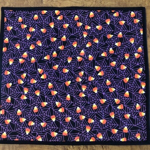 Op de afbeelding: Een zwarte en paarse stoffen placemat met een patroon van witte spinnenwebben en oranje en witte candy corn.