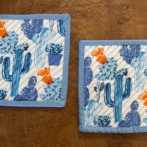 May include: Two blue and white quilted pot holders with a cactus pattern. The pot holders have a blue border.